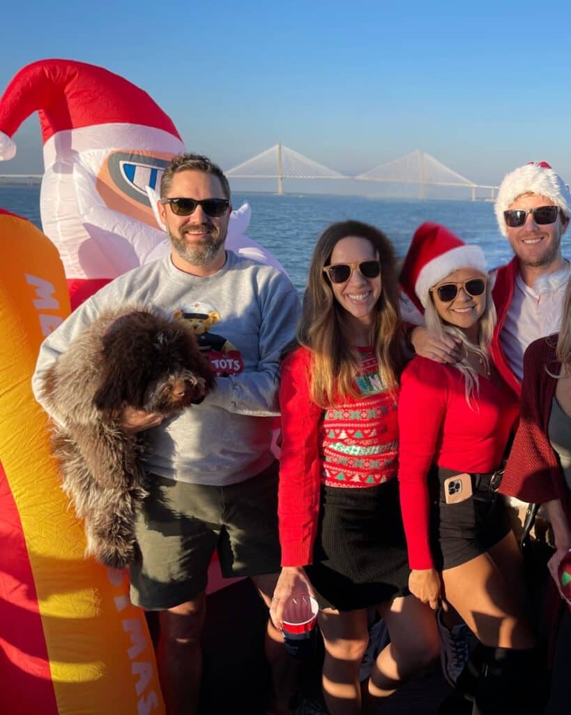 Friends in Christmas outfits posing with a Santa inflatable on a holiday-themed boat cruise in Charleston Harbor.