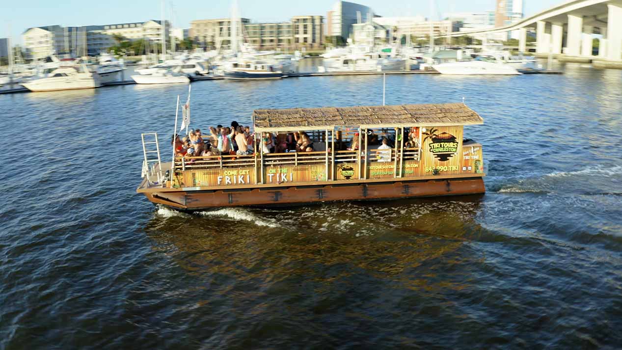BOAT-TOUR-CHARLESTON-2 The Friki Tiki boat navigating a scenic harbor boat tour in Charleston, SC, showcasing its tropical roof and spacious passenger deck.
